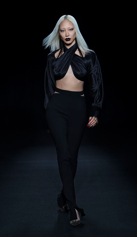 A model with straight, long platinum blonde hair walks confidently down a dark runway, wearing a stylish black crop top and high-waisted black pants. The outfit features cut-out details, and she complements her look with bold dark lipstick and striking accessories.