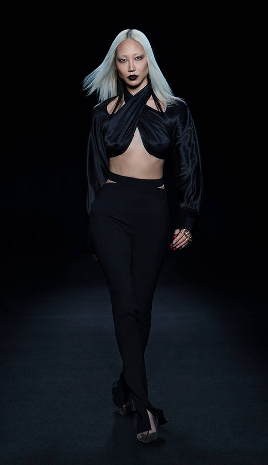 A model with straight, long platinum blonde hair walks confidently down a dark runway, wearing a stylish black crop top and high-waisted black pants. The outfit features cut-out details, and she complements her look with bold dark lipstick and striking accessories.