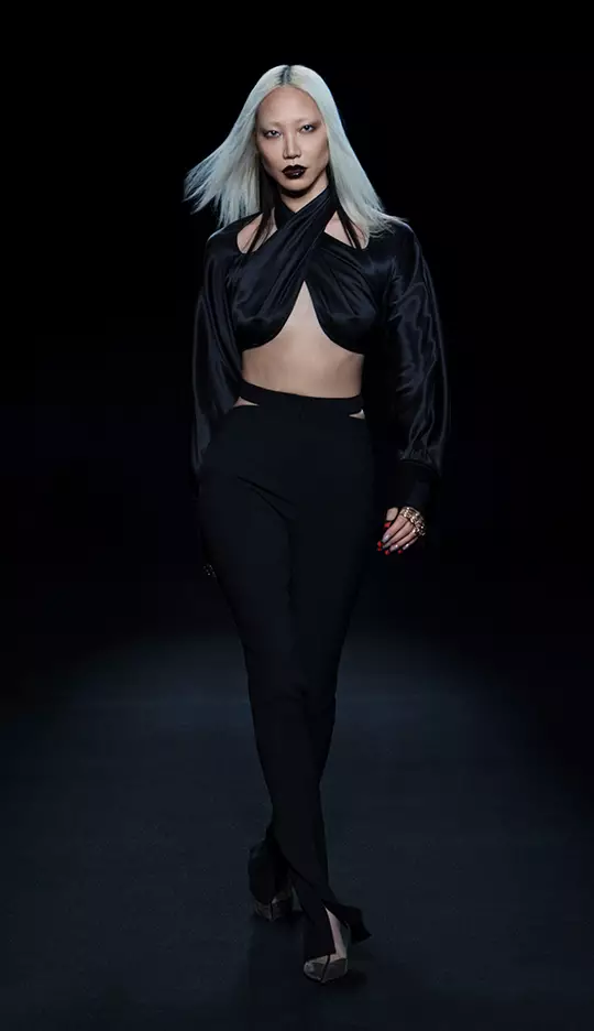 A model with straight, long platinum blonde hair walks confidently down a dark runway, wearing a stylish black crop top and high-waisted black pants. The outfit features cut-out details, and she complements her look with bold dark lipstick and striking accessories.