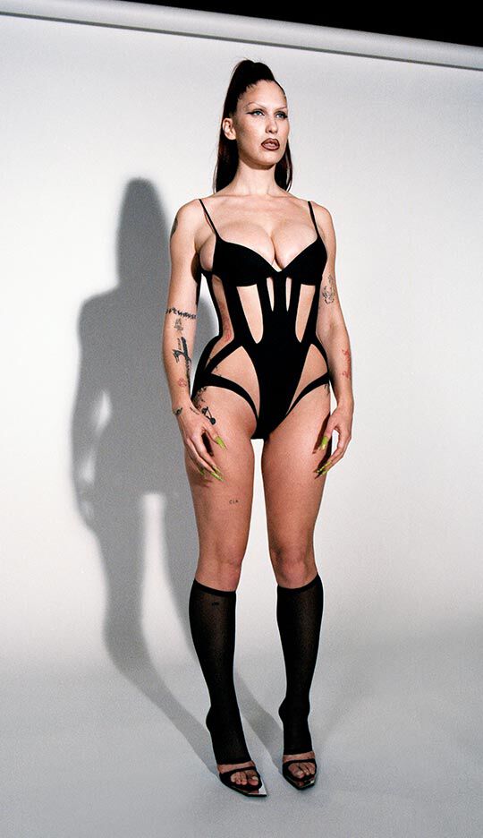 A person stands against a neutral background, wearing a stylish black bodysuit with cut-out designs, high heels, and long, elegantly manicured nails. Their hair is styled up, and they have bold makeup.