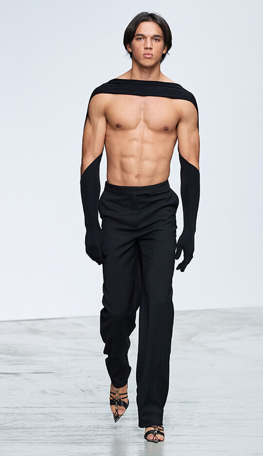 A muscular male model walks down the runway wearing black pants and a unique top that features long sleeves extending over his shoulders, showcasing an athletic physique.