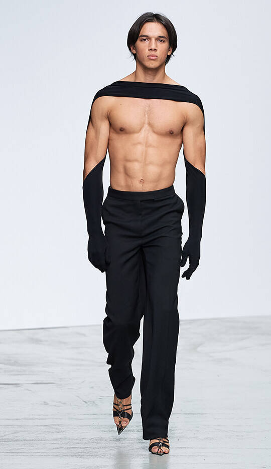 A muscular male model walks down the runway wearing black pants and a unique top that features long sleeves extending over his shoulders, showcasing an athletic physique.