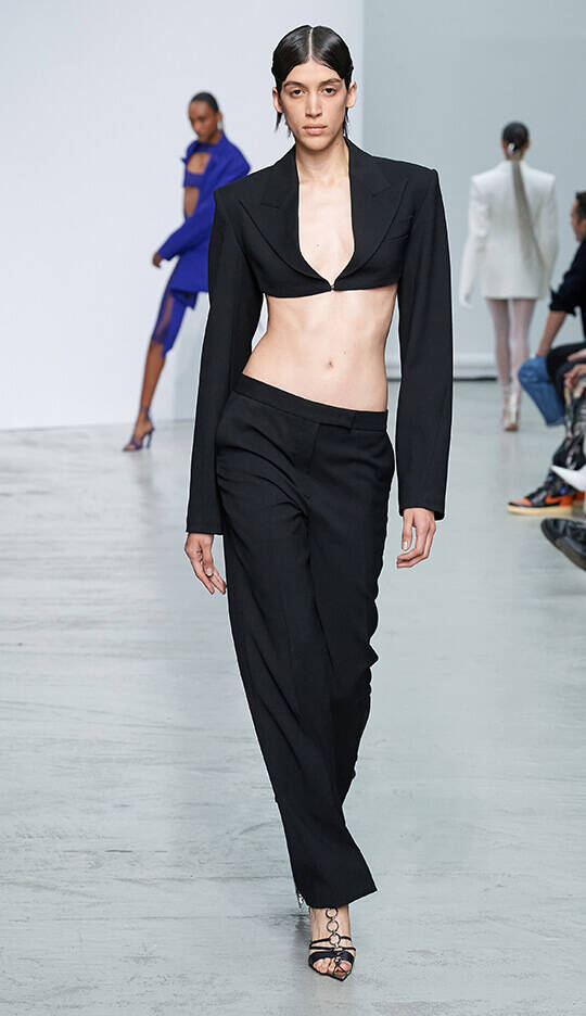 A model walks confidently down the runway wearing a black cropped blazer and wide-leg trousers, showcasing a modern and edgy fashion look. In the background, another model poses in a blue outfit.