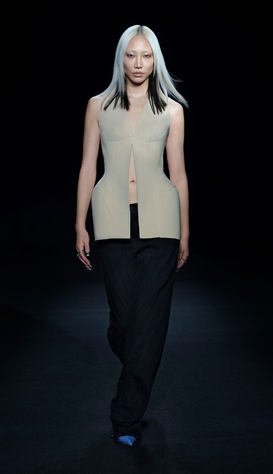 A model walks down the runway wearing a modern, sleeveless beige top with a unique cut, paired with black, wide-legged pants. The background is dark, highlighting the striking outfit and the model's sleek, straight hair.