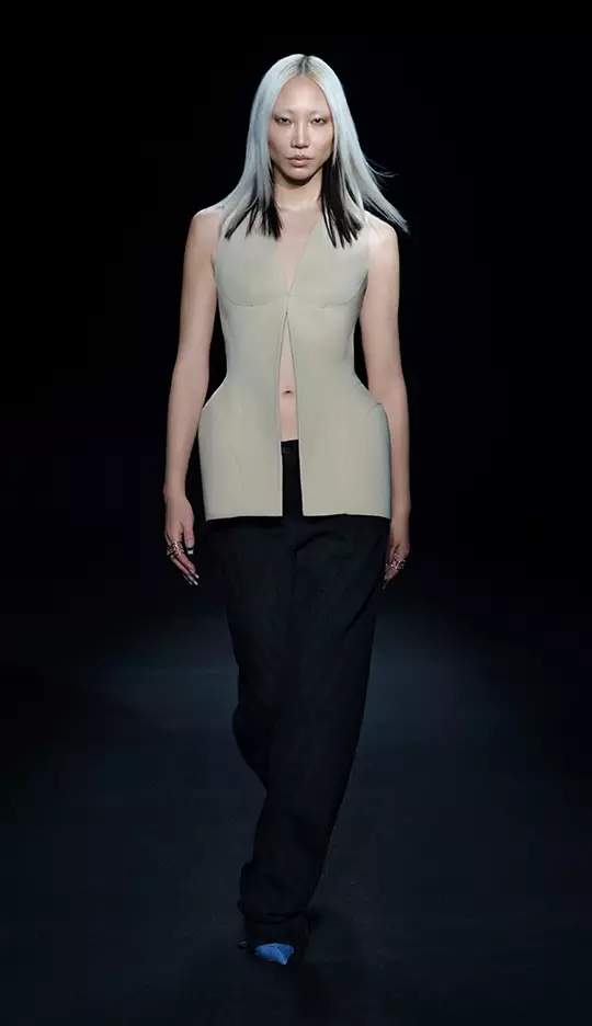 A model walks down the runway wearing a modern, sleeveless beige top with a unique cut, paired with black, wide-legged pants. The background is dark, highlighting the striking outfit and the model's sleek, straight hair.