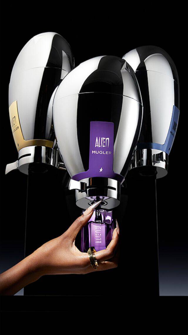 A hand holding a purple perfume bottle is positioned below three shiny, stylized perfume containers on a sleek black background.