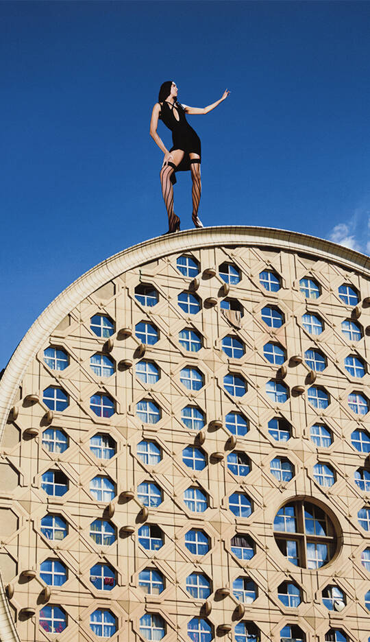 A person in a black outfit stands on top of a circular building with a patterned facade and multiple windows, under a clear blue sky.