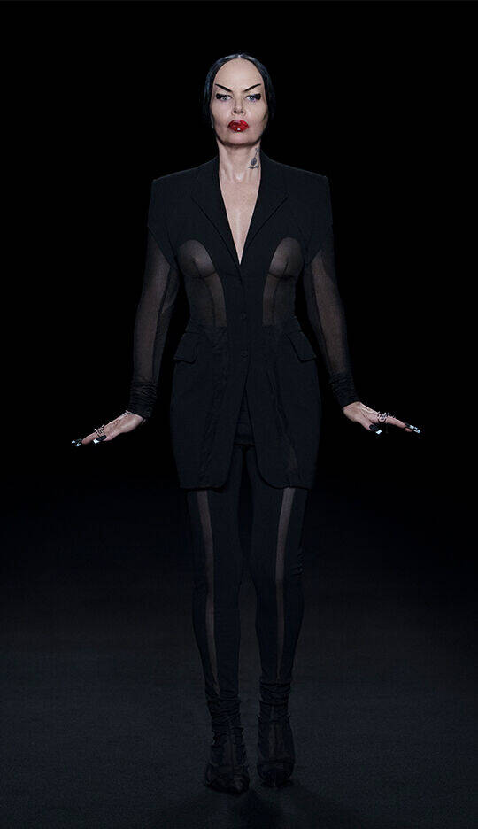 A woman stands confidently in a dark, minimalist setting, wearing a black outfit with sheer panels and a tailored blazer. She has bold red lipstick and holds her hands out to the sides, exuding an edgy, fashion-forward vibe.