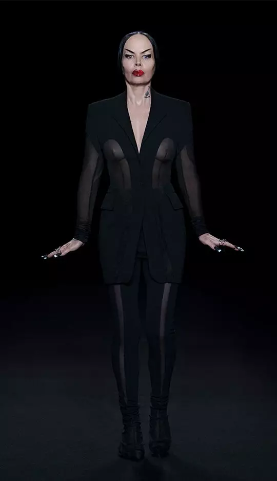 A woman stands confidently in a dark, minimalist setting, wearing a black outfit with sheer panels and a tailored blazer. She has bold red lipstick and holds her hands out to the sides, exuding an edgy, fashion-forward vibe.