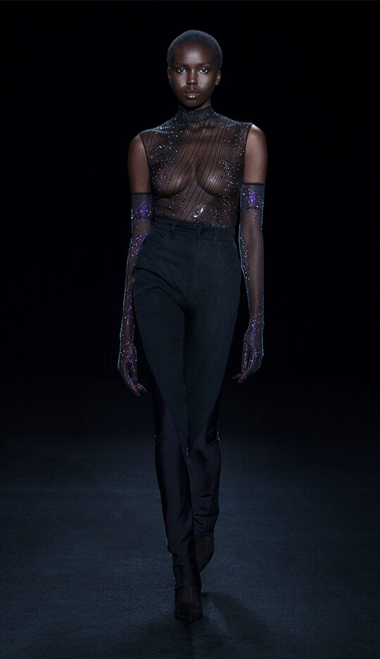 A model walks down a dark runway wearing a sheer, embellished top and high-waisted black pants, complemented by long, sparkly gloves. The overall look is elegant and striking against the dark background.