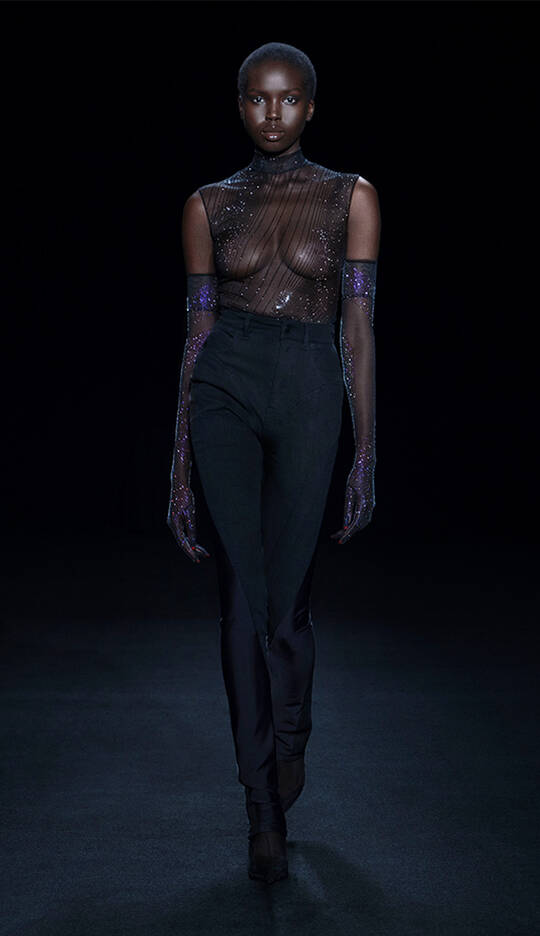 A model walks down a dark runway wearing a sheer, embellished top and high-waisted black pants, complemented by long, sparkly gloves. The overall look is elegant and striking against the dark background.
