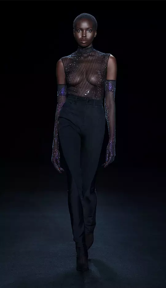 A model walks down a dark runway wearing a sheer, embellished top and high-waisted black pants, complemented by long, sparkly gloves. The overall look is elegant and striking against the dark background.