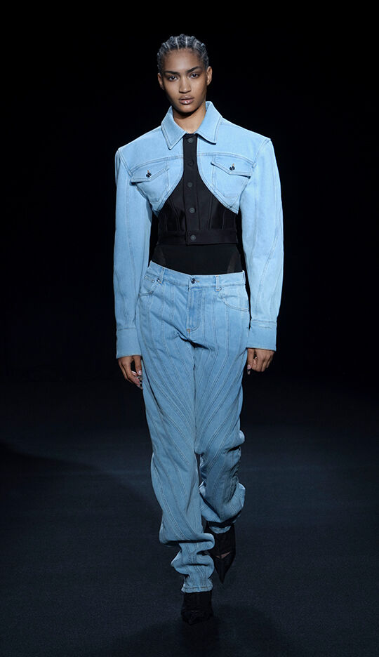 A model walks down the runway wearing a light blue oversized denim jacket paired with a black fitted top and light blue striped jeans. The backdrop is dark, emphasizing the outfit.