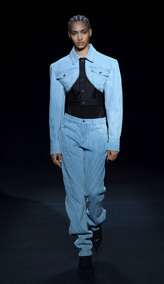A model walks down the runway wearing a light blue oversized denim jacket paired with a black fitted top and light blue striped jeans. The backdrop is dark, emphasizing the outfit.