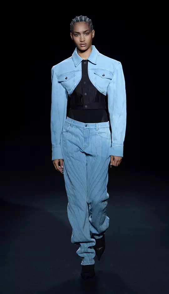 A model walks down the runway wearing a light blue oversized denim jacket paired with a black fitted top and light blue striped jeans. The backdrop is dark, emphasizing the outfit.