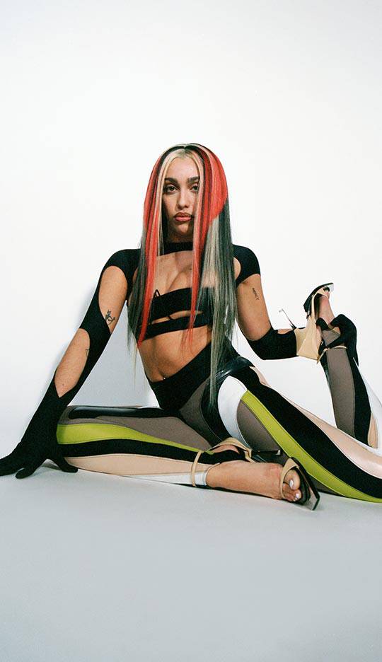 A person sits on the floor in a stylish pose, wearing a black and colorful outfit that features a crop top and form-fitting leggings. Their long hair is styled in a noticeable red and gray combination, and they are holding a high-heeled shoe in one hand. The background is plain and minimalistic.