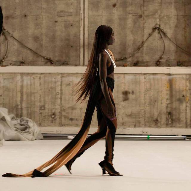 A model walks down a runway in a striking outfit with a long, flowing train, set against a minimalist backdrop of a photo studio.