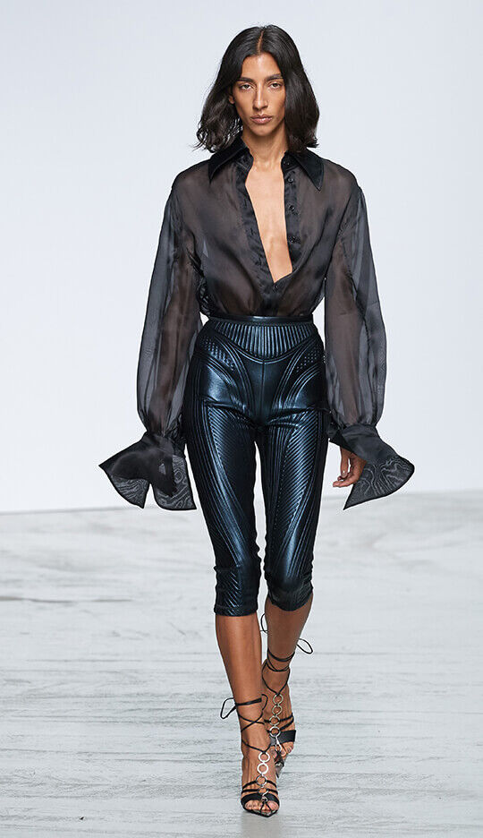 A model walks down the runway wearing a sheer black blouse with voluminous sleeves paired with shiny fitted pants and heeled sandals. The background is simple and white, emphasizing the outfit.