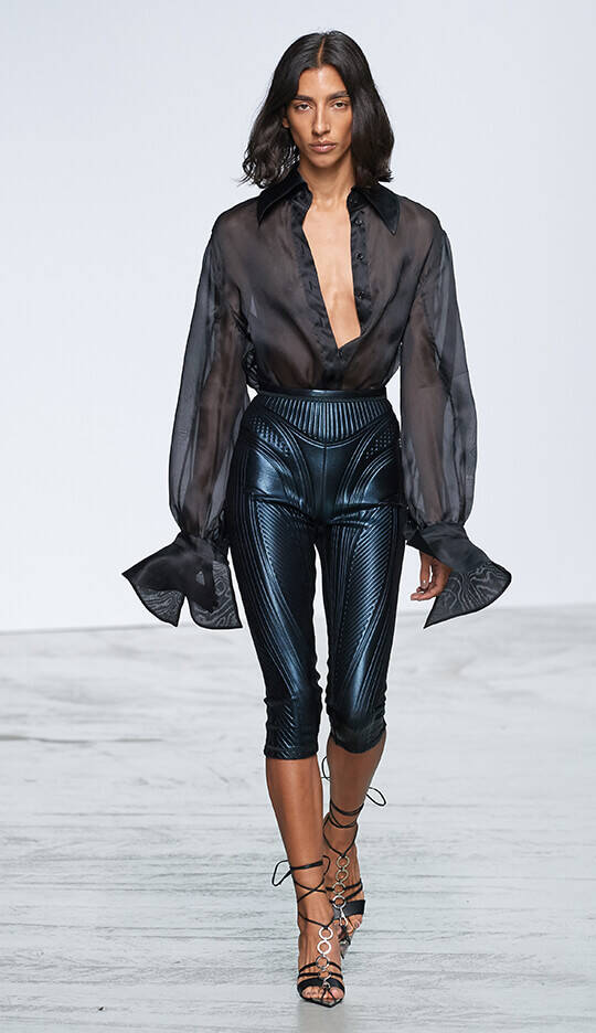 A model walks down the runway wearing a sheer black blouse with voluminous sleeves paired with shiny fitted pants and heeled sandals. The background is simple and white, emphasizing the outfit.