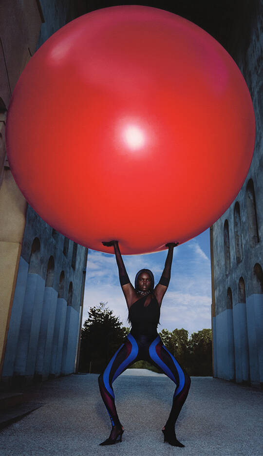 A person in a black outfit and colorful leggings lifts a large red ball above their head against a backdrop of a blue sky and architectural structures.