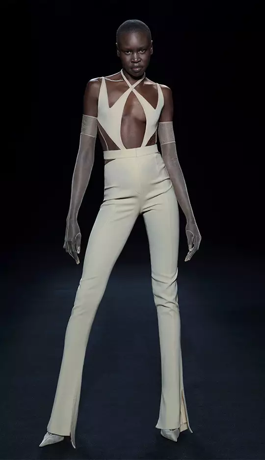 A model poses in a minimalist, form-fitting beige outfit with a unique cutout design, complemented by sheer gloves and heels, set against a dark background.
