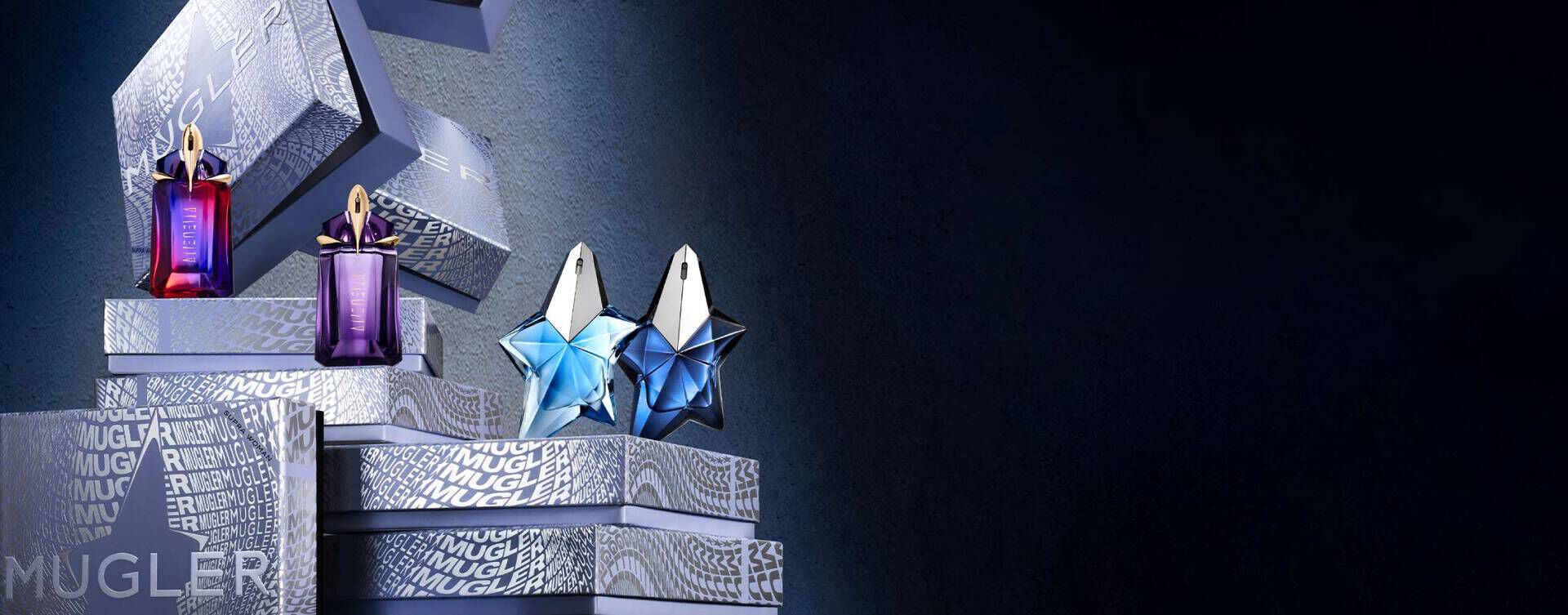 An array of Mugler fragrances, including Alien and star-shaped Angel bottles, displayed on branded gift boxes against a dark background.