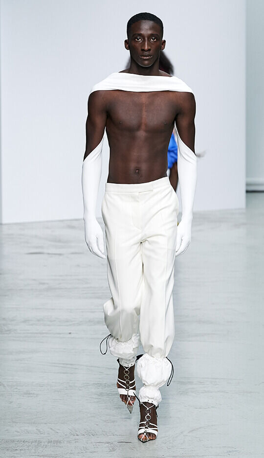 A model walks down the runway wearing a stylish ensemble, featuring a bare chest, high-waisted white trousers, and long white gloves, paired with unique strappy shoes. The backdrop is minimalist, emphasizing the fashion design.
