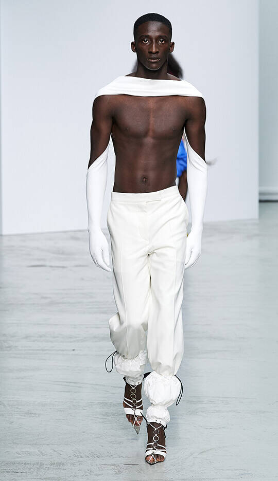 A model walks down the runway wearing a stylish ensemble, featuring a bare chest, high-waisted white trousers, and long white gloves, paired with unique strappy shoes. The backdrop is minimalist, emphasizing the fashion design.