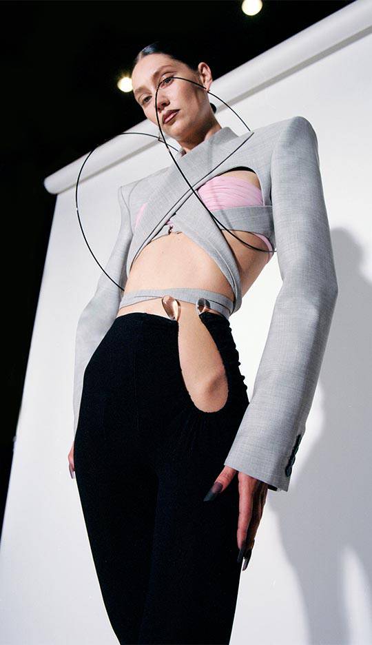 A model poses in a futuristic outfit featuring a structured gray blazer with unique cutouts, paired with black pants that have distinctive openings. The setting is a photoshoot with a plain backdrop.