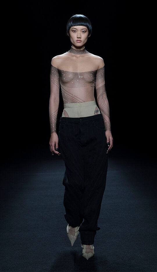 A model walks down a runway wearing a sheer off-the-shoulder top with shimmering designs and loose black pants, showcasing a contemporary fashion look. The background is dark, highlighting the outfit.