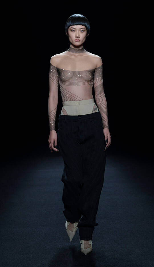 A model walks down a runway wearing a sheer off-the-shoulder top with shimmering designs and loose black pants, showcasing a contemporary fashion look. The background is dark, highlighting the outfit.