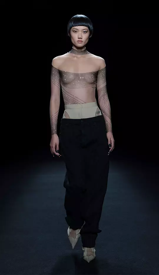 A model walks down a runway wearing a sheer off-the-shoulder top with shimmering designs and loose black pants, showcasing a contemporary fashion look. The background is dark, highlighting the outfit.