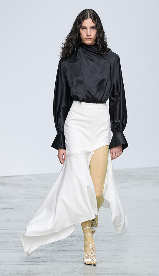A model walks down the runway wearing a stylish black satin top paired with a flowing white skirt and yellow-accented sheer tights, showcasing a modern fashion look.