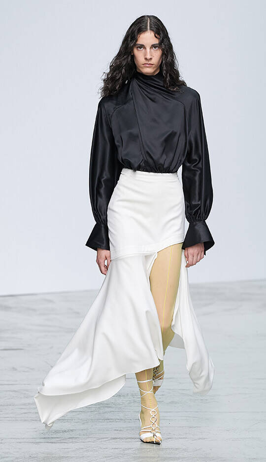 A model walks down the runway wearing a stylish black satin top paired with a flowing white skirt and yellow-accented sheer tights, showcasing a modern fashion look.