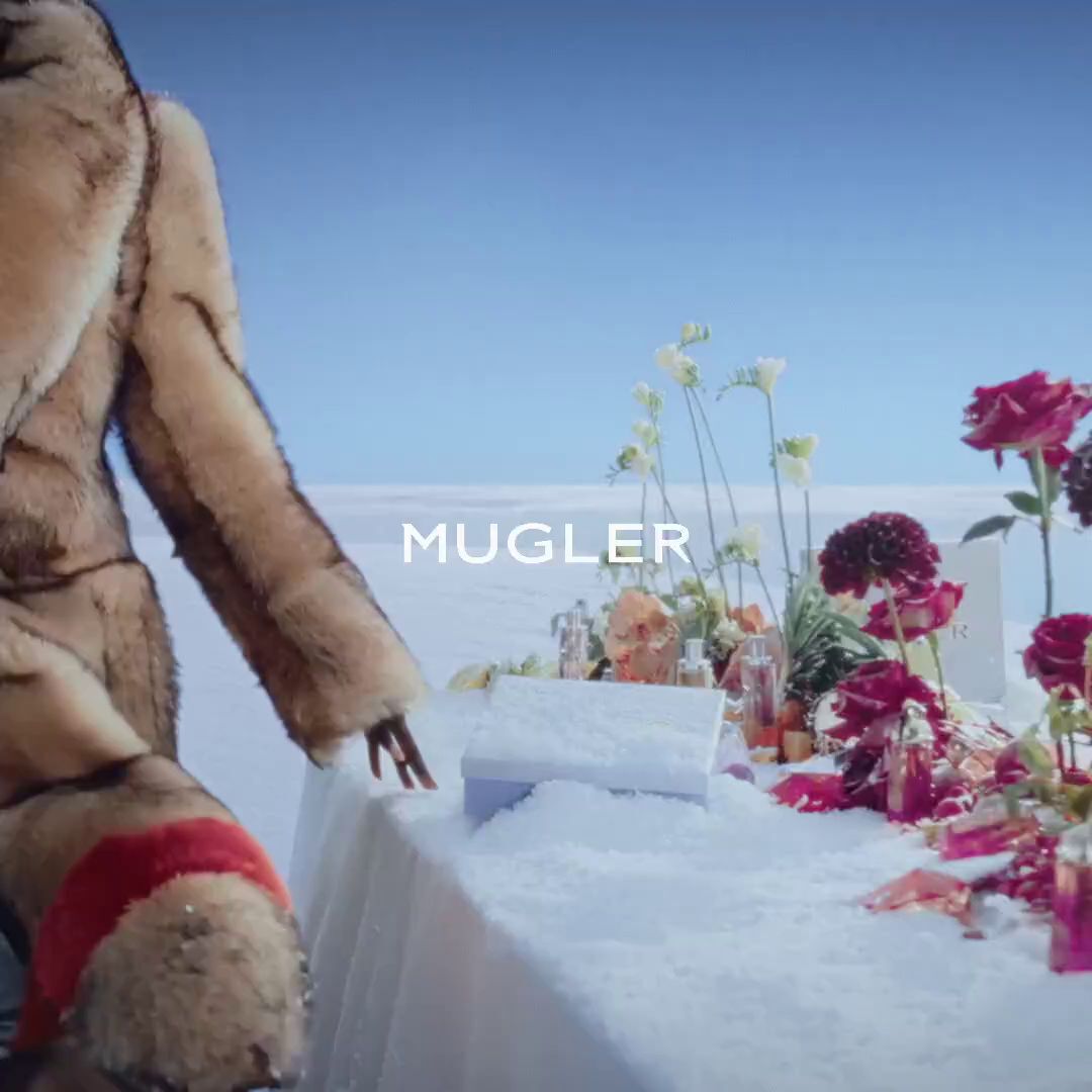 A Mugler campaign video showing an icy blue gift set buried in snow. A hand opens a gift set to reveal the iconic blue star-shaped Angel Eau de Parfum bottle lying in the snow. The video ends with the blue star bottle standing upright in front of white Mugler gift boxes against a blue sky.