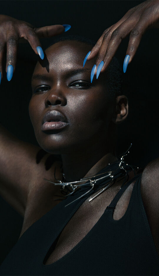 A close-up portrait of a dark-skinned person with a shaved head, striking a pose with hands raised and long blue-painted nails. They are wearing a black top with a bold, spiked necklace, and the lighting creates dramatic shadows on their face.
