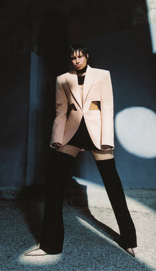 A person poses dramatically in a stylish outfit featuring a tailored beige blazer with cutouts, paired with black pants that create an elongated silhouette. The background is a mix of light and shadow, adding depth to the striking look.