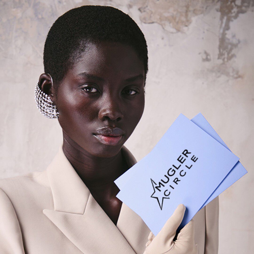 Model in beige blazer holding two light purple Mugler cards.