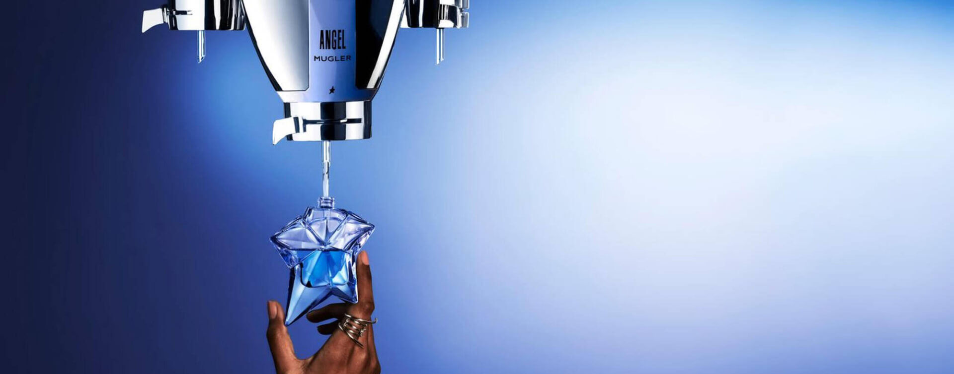 A hand reaching to catch a blue star-shaped perfume bottle as liquid is dispensed from a sleek silver perfume dispenser against a gradient blue background.