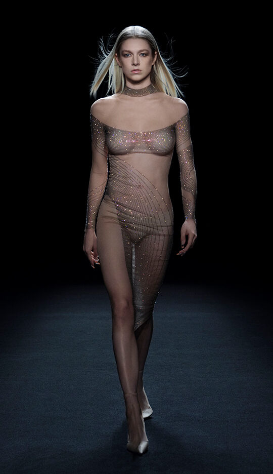 A model walks the runway wearing a sheer, glittering dress that features a delicate lattice design, in a darkened setting.