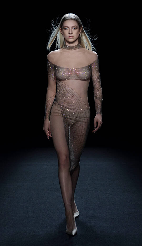 A model walks the runway wearing a sheer, glittering dress that features a delicate lattice design, in a darkened setting.