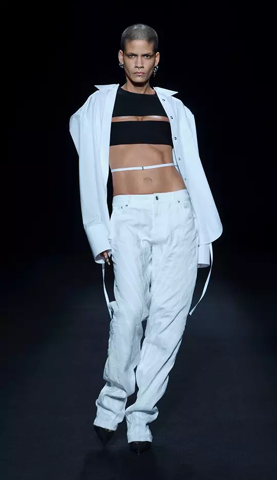 A model walks down the runway wearing a black cropped top with horizontal cutouts and a large white oversized shirt, paired with white wide-legged pants and black heels, against a dark backdrop.