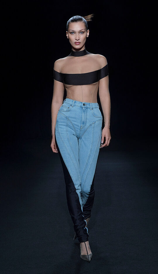 A model walks down a runway wearing a daring outfit that features a sheer black top and unique, form-fitting jeans. The background is dark, highlighting the fashion design.