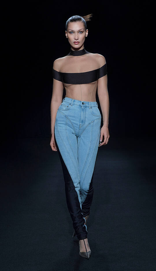 A model walks down a runway wearing a daring outfit that features a sheer black top and unique, form-fitting jeans. The background is dark, highlighting the fashion design.