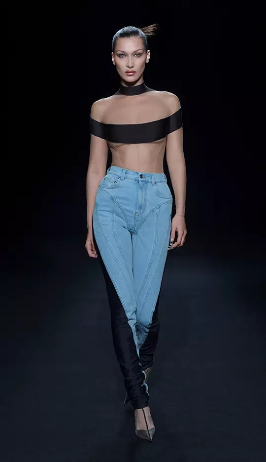 A model walks down a runway wearing a daring outfit that features a sheer black top and unique, form-fitting jeans. The background is dark, highlighting the fashion design.