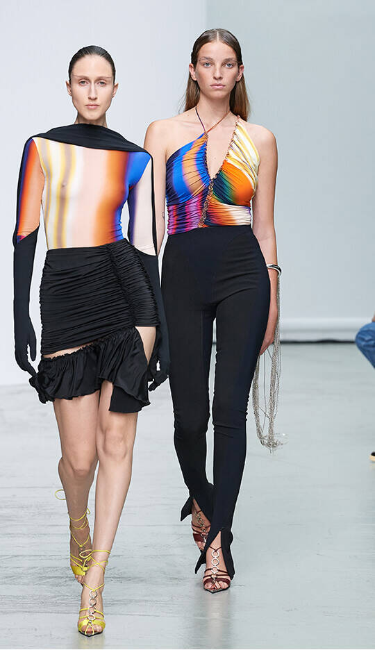 Two models are walking down a runway, showcasing vibrant, colorful outfits. One model wears a long-sleeve top with abstract patterns and a draped, asymmetrical skirt, while the other model sports a sleeveless, form-fitting top and sleek black pants. The backdrop is simple and modern.