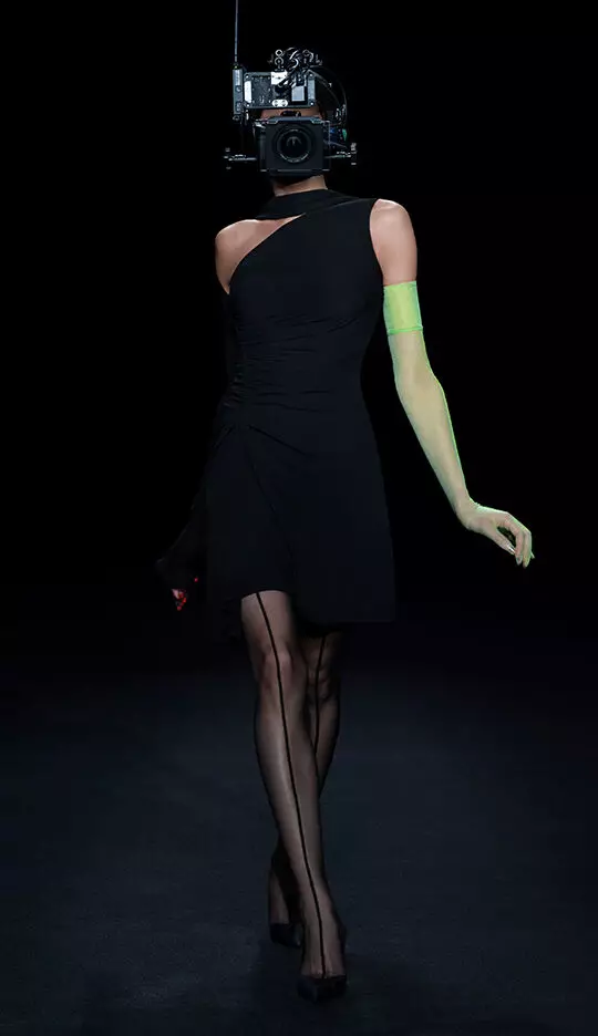 A model wears a sleek black dress with an asymmetrical cut, paired with green arm accessories and fishnet stockings, while a vintage camera is attached to her head, creating a surreal fashion statement against a dark background.