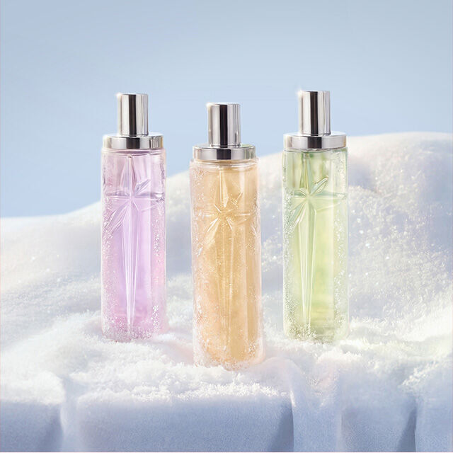 Three tall, cylindrical Mugler fragrance bottles in translucent shades of pink, orange, and light green standing upright in a row in the snow. The new Starlicious collection. text reads "gifts under $50"