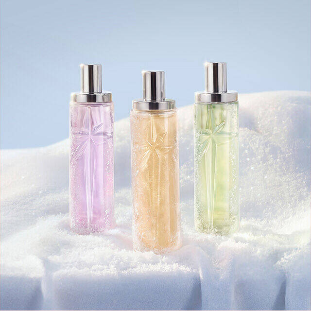 Three tall, cylindrical Mugler fragrance bottles in translucent shades of pink, orange, and light green standing upright in a row in the snow. The new Starlicious collection. text reads "gifts under $50"