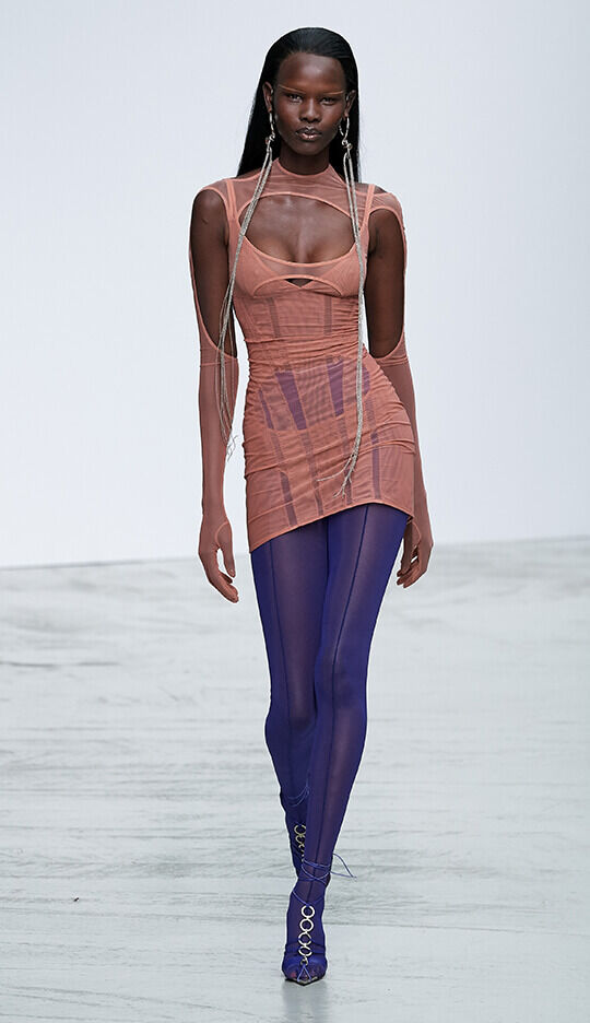 A model walks down a minimalist runway wearing a form-fitting, layered outfit with a combination of soft pink and bright blue tights, showcasing a modern and artistic fashion design.